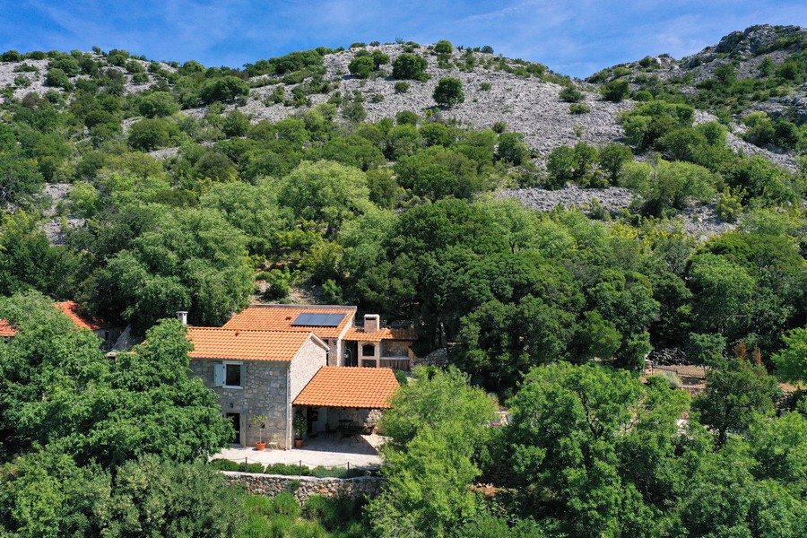 Vogelperspektive auf ein kroatisches Steinhaus zum Verkauf, eingebettet in mediterranes Grün mit sichtbaren Gebirgsformationen im Hintergrund.