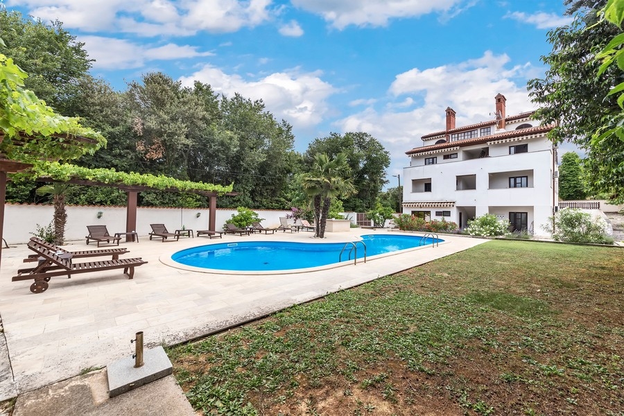 Ein moderner Poolbereich mit Liegestühlen und einem großen weißen Haus im Hintergrund, umgeben von Bäumen